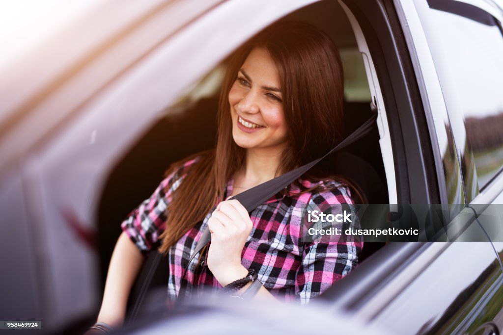 Young joyful beautiful girl is buckling up in a drivers seat of her car. Driving Stock Photo