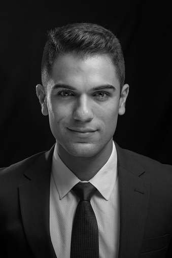 Young business man poses for a studio portrait. He is his twenties and has black hair and dark brown eyes. He is wearing a formal outfit with neck tie, coat and white shirt. He is looking at the camera. This is a monochrome image.