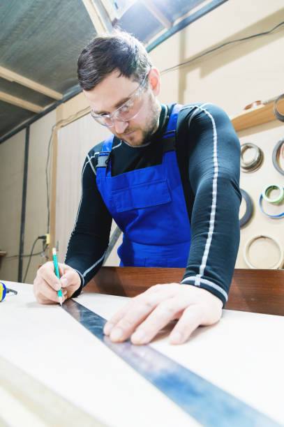 istock A bearded tired handsome carpenter on a table with a pencil draws a sign on the board. At the workplace there is an ordinary master dressed in blue cambenizon with a cruel haircut and saved glasses. The concept of garage furniture production 958510518