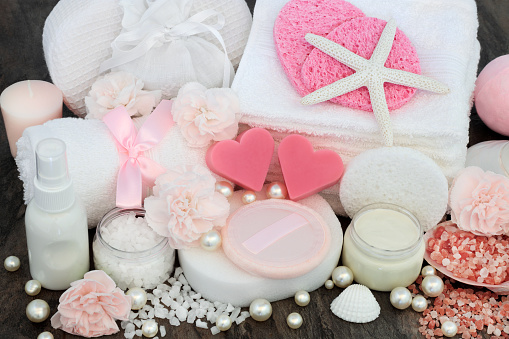 Skin care and ex foliation beauty treatment with himalayan ex foliating salts, heart shaped pink soaps, sponges, face cloths, body lotion, moisturising cream, decorative shells and pearls on marble background.
