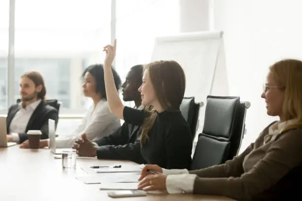 Photo of Smiling curious businesswoman raising hand at group meeting voting as volunteer