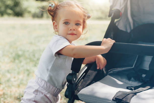Baby girl standing near a baby carriage A little girl is rolling baby carriage in the park. Child in the park playing with pram babyhood stock pictures, royalty-free photos & images