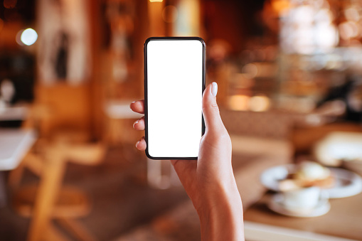 Girls hands holding white screen smartphone on blurred abstract brown cafe background.