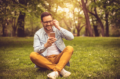 Young happy man sitting on grass and listening music on smart phone.
