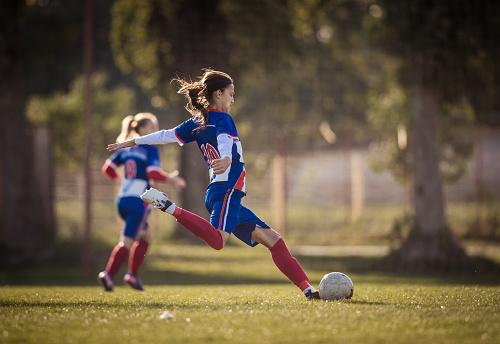 Teenage girl running with soccer ball on a playing field during the match and about to kick the ball.