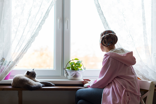 Young adult female cancer patient wearing headscarf and bathrobe sitting in the kitchen with her pet cat, looking out window.