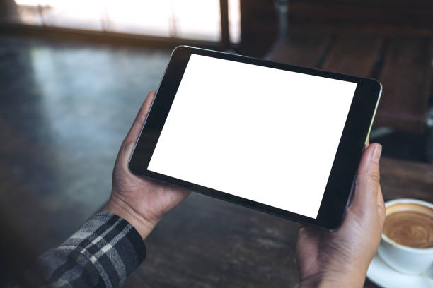 istock Mockup image of hands holding black tablet pc with white blank screen and coffee cup on table background 958707256