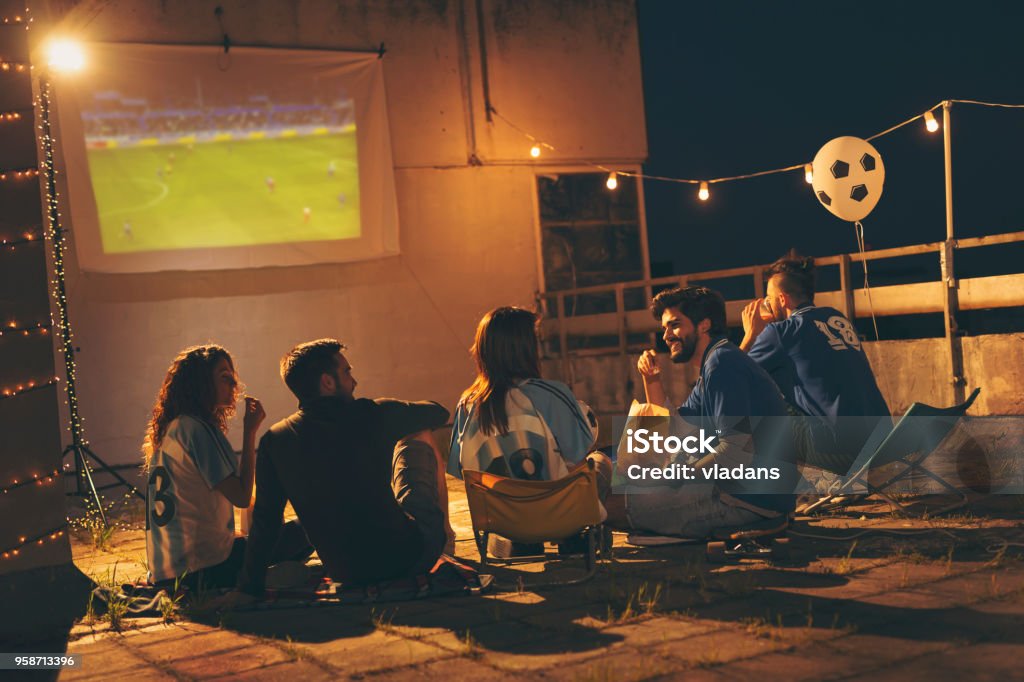 Group of friends watching football on a building rooftop Group of young friends having fun while watching a football match on a building rooftop. Focus on the guy eating popcorn Watching Stock Photo