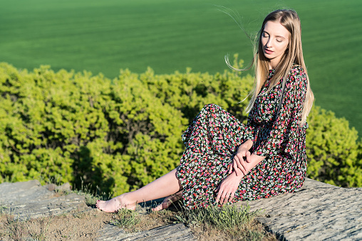 Romantic picture of young pretty model in dress sitting on rock with beautiful green field in background