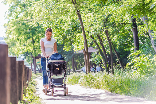 Family, child and parenthood concept - Front view of happy mother walking with baby stroller in park during bright summer day. Photo of Young Caucasian mother pushing pram in park on sunny day.   Full length of Beautiful young Female pushing baby carriage in nature.