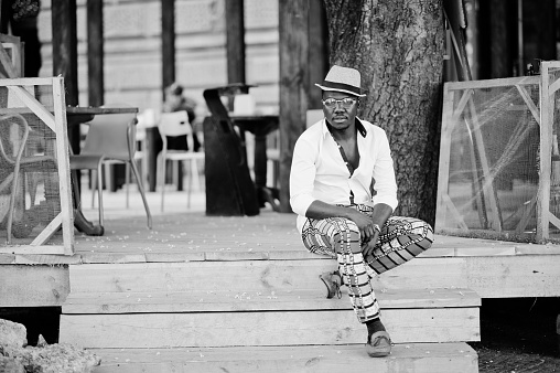Stylish african american man in white shirt and colored pants with hat and glasses posed outdoor. Black fashionable model boy.