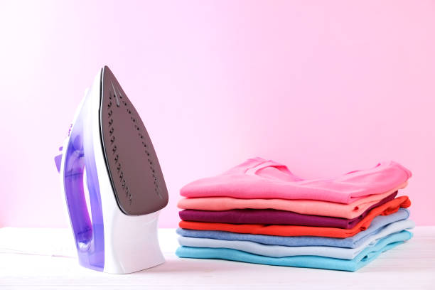 Neat stack of clean freshly laundered clothes and electric iron on pressboard. Pile of t-shirts and sweaters on white wooden ironing board, pink wall background. Copy space, close up, top view.
