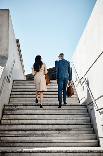 Rear view of business people moving up stairs. Male and female professionals walking on sunny day. Full length of colleagues are in long coat and suit.