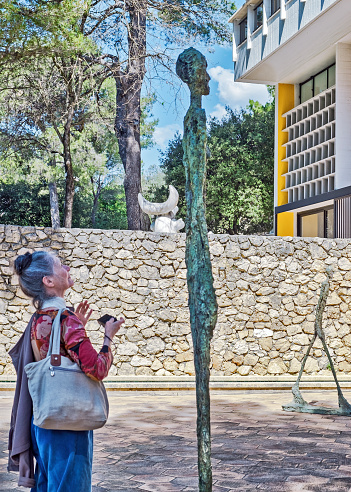 Saint-Paul-de-Vence, France - April 17, 2018: A snapshot of a mature woman holding a camera viewing a sculpture by Alberto Giacometti in the courtyard of the Foundation Maeght, an art institution in Saint-Paul-de-Vence, France. In the background sculptures by Alberto Giacometti, Joan Miró and modern architecture.