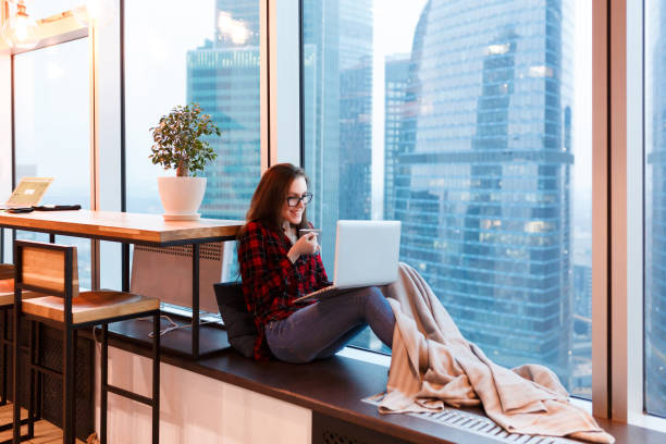 istock Cute young woman outsourcing designer working in creative studio sitting under blanket with mug of coffee and working at the computer on background of large window overlooking the skyscrapers 958793828