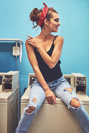 Shot of an attractive young woman seated on a washing machine while waiting for the washing to be washed inside of a laundry room