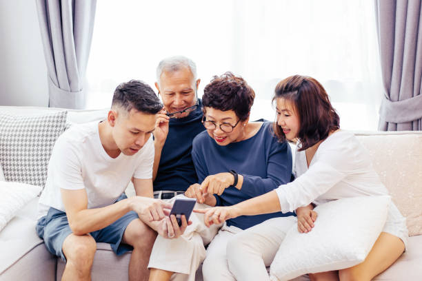 família asiática com filhos adultos e sênior pais usando um telefone celular e relaxando em um sofá em casa juntos - digital tablet family father offspring - fotografias e filmes do acervo