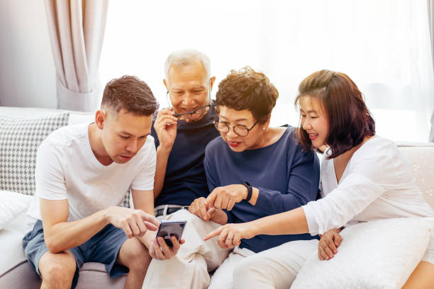 asian family with adult children and senior parents using a mobile phone and relaxing on a sofa at home together - father digital tablet asian ethnicity daughter imagens e fotografias de stock