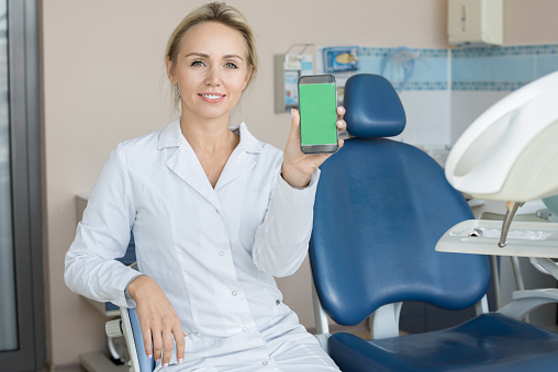 Portrait of beautiful female dentist posing in dental clinic holding smartphone with green screen and presenting mobile app or website to camera, copy space