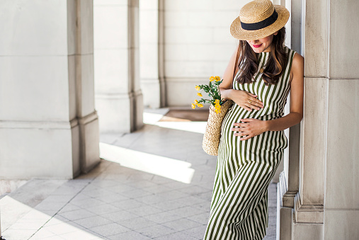 A pregnant woman standing with flowers wearing a green and white striped dress and a straw hat.