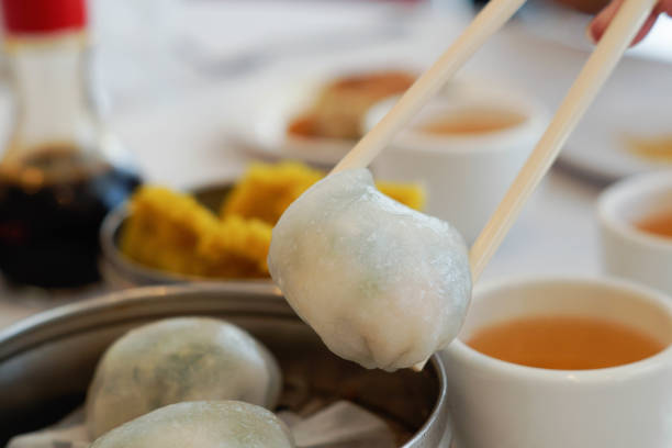 Dim sum table with tea and soy sauce in the background.  A dim sum crystal dumpling that has shrimp, pork and garlic chives is being pick up with chopsticks.