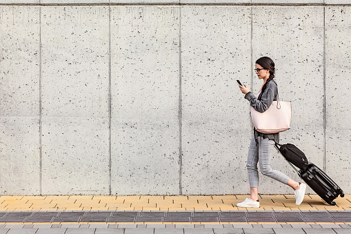 Attractive young woman using her smart phone walking beside the concrete wall. She is pulling a small wheeled luggage with a briefcase on it and it seems that she is in a good mood. The shot is executed with available natural light, and the copy space has been left.