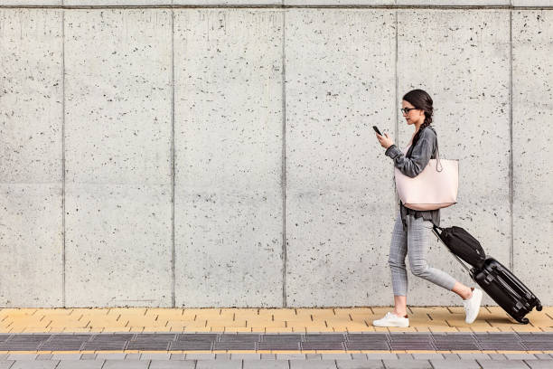 Young woman using her smart phone walking beside the concrete wall and pulling a small wheeled luggage with a briefcase on it stock photo