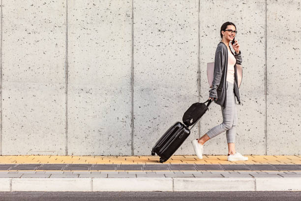 istock Young woman using her smart phone walking beside the concrete wall and pulling a small wheeled luggage with a briefcase on it 958923500