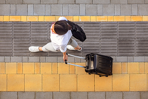 High angle view of attractive young woman walking on a sidewalk and pulling a small wheeled luggage with a briefcase on it. It seems that she is in a good mood. The shot is executed with available natural light, and the copy space has been left.