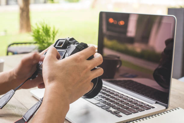istock Man hand holding camera and using laptop computer in coffee shop. 958928264