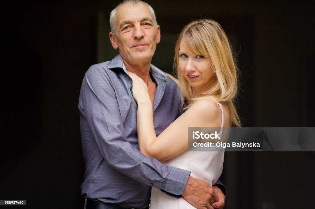 Couple with Age Difference Hugging on Black Background. Attractive Young Woman in Dress and Senior Man in Blue Shirt Embracing and Looking at the Camera Couple with Age Difference Hugging on Black Background. Attractive Young Woman in Dress and Senior Man in Blue Shirt Embracing and Looking at the Camera. Aging Process Stock Photo