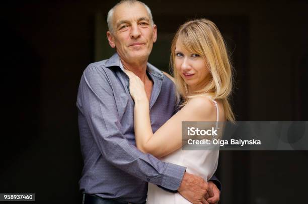 Couple With Age Difference Hugging On Black Background Attractive Young Woman In Dress And Senior Man In Blue Shirt Embracing And Looking At The Camera Stock Photo - Download Image Now
