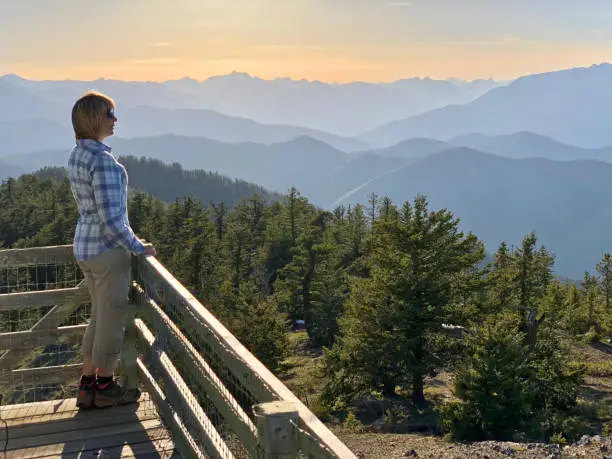 Photo of Ordinary middle age woman hiker on the balcony of fire watch tower enjoying beautiful views of mountains at sunset.