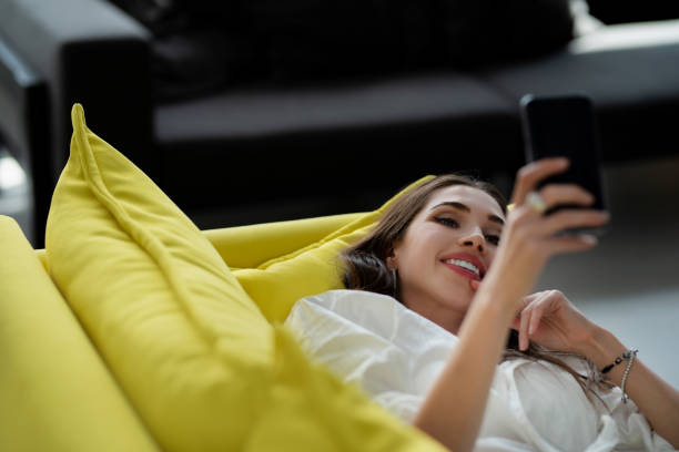 una bella giovane donna sdraiata su un divano, sta usando un'applicazione nel suo dispositivo smartphone - living room elegance women long hair foto e immagini stock