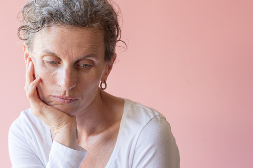 Head and shoulders view of middle aged woman with head resting on chin looking down against pink background (selective focus)