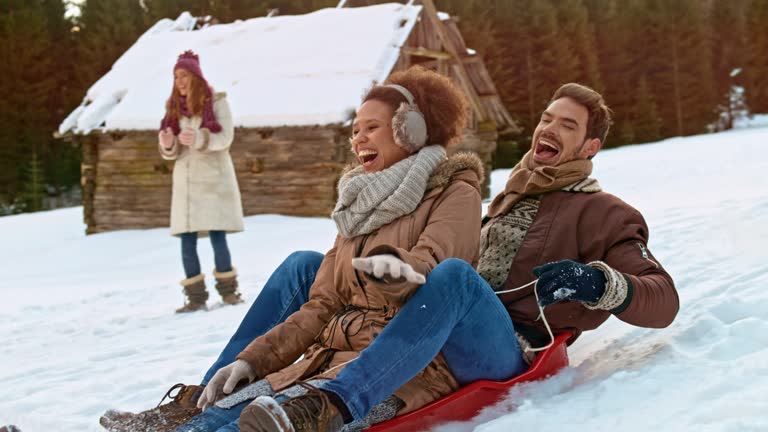 Slow motion wide handheld shot of a man and a woman laughing while gliding down the hill in a snow sledge after being pushed by their male friend. Shot in Slovenia.