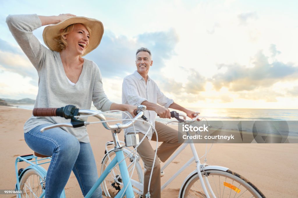 Mature couple cycling on the beach at sunset or sunrise. Mature couple cycling on the beach at sunset or sunrise. They are laughing and having fun. They are casually dressed. Could be a retirement vacation. Cycling Stock Photo
