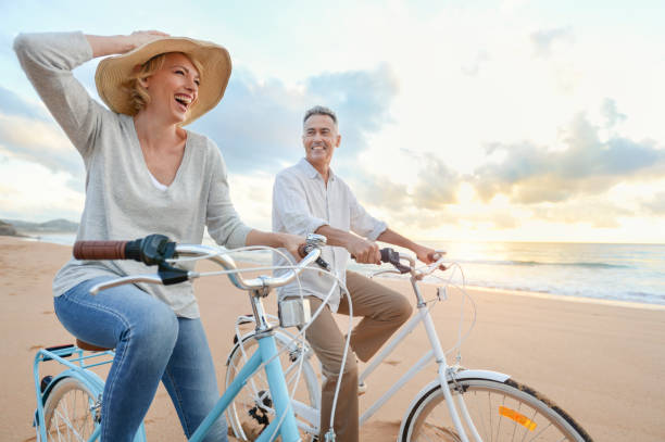 Mature couple cycling on the beach at sunset or sunrise. Mature couple cycling on the beach at sunset or sunrise. They are laughing and having fun. They are casually dressed. Could be a retirement vacation. beach lifestyle stock pictures, royalty-free photos & images