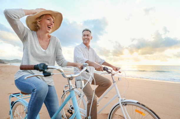 dojrzała para na rowerze na plaży o zachodzie słońca lub wschodzie słońca. - married couple mature adult outdoors zdjęcia i obrazy z banku zdjęć