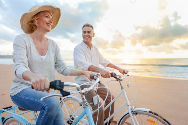 coppia matura in bicicletta sulla spiaggia al tramonto o all'alba. - mature adult bicycle senior adult heterosexual couple foto e immagini stock