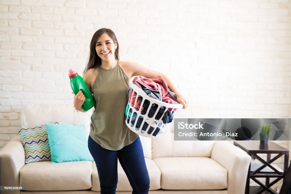 Beautiful young woman is holding a fabric softener and laundry basket filled with clothes Front view of a young cheerful housewife holding basket with dirty clothes for wash and detergent bottle Laundry Stock Photo