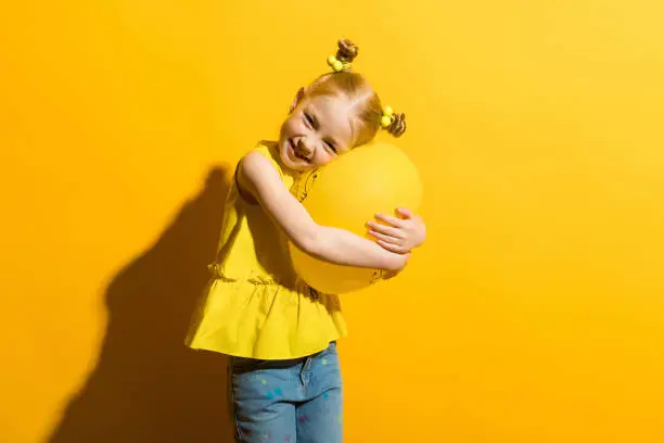 Photo of Girl with red hair on a yellow background. A girl is hugging a yellow balloon.