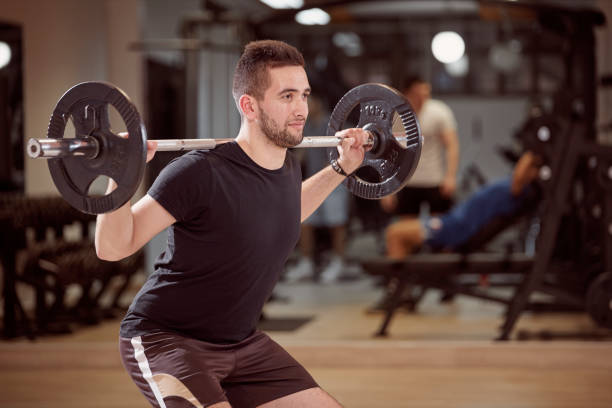istock upper body shot, squat exercise, young man holding barbell with weights on back shoulders. Unrecognizable people behind in gym (out of focus) 959027538