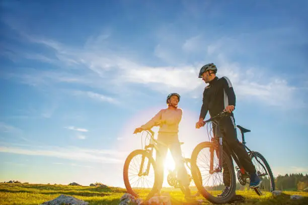 Cyclist couple with mountain bikes standing on the hill under the evening sky and enjoying bright sun at the sunset. Below is a city in the distance.