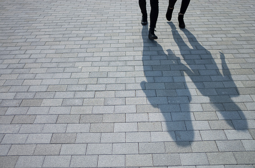 Two women are walking on the street.\nThe shadows are growing from the feet of them.