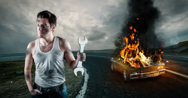 Man holding a wrench with his car on fire in the background. Needs help to repair the car. Car insurance concept.