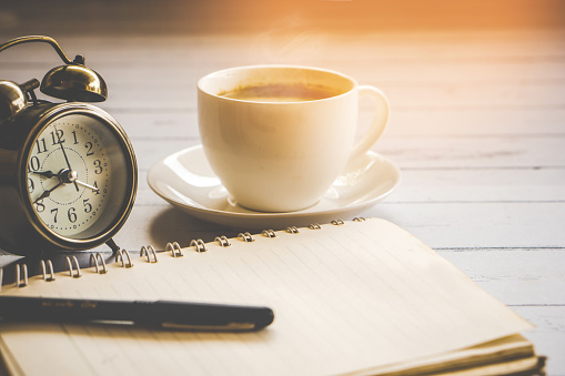 cup of hot coffee ,clock,notebook and pen on wooden table with sunlight morning background