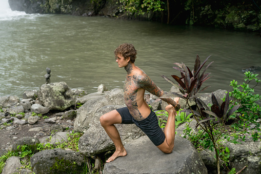 tattooed man practicing yoga on rock with river and green plants on background, Bali, Indonesia