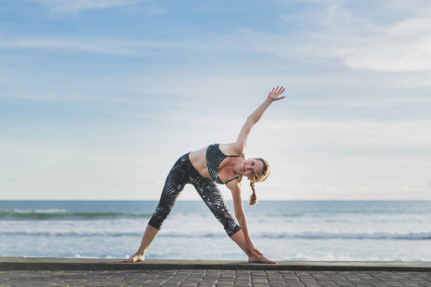 istock woman practicing yoga in Extended Triangle Pose with ocean and blue sky on background, Bali, Indonesia 959109350