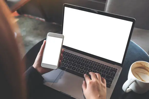 Photo of Top view mockup image of hands holding blank mobile phone while using laptop with blank white desktop screen on table